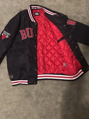Chicago Bulls bomberjacka NBA - Snygg svart bomberjacka från NBA med Chicago Bulls-logga och broderade detaljer i rött och vitt. Jackan har röd quiltad insida, randiga muddar och tryckknappar framtill. Broderad text på bröstet och ärmen samt nummerdetalj på ärmen.