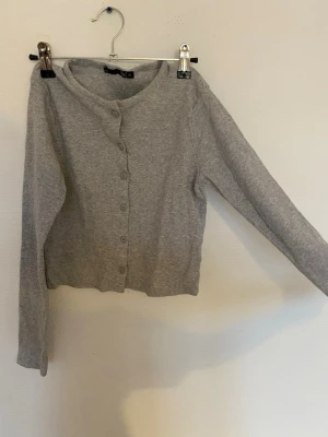 Grå ribbad kofta med knappar - Mysig grå kofta med ribbad struktur och knäppning framtill. Modellen är kortare och har långa ärmar. Perfekt att slänga över en topp för en enkel och chill look.