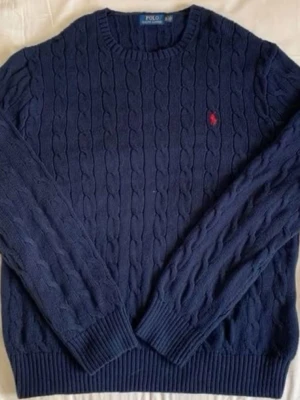 Mörkblå kabelstickad tröja Polo Ralph Lauren - Kabelstickad mörkblå tröja från Polo Ralph Lauren med rund halsringning och den klassiska röda logotypen broderad på bröstet. Tröjan har ribbade muddar vid ärmslut och nederkant. Perfekt för dig som gillar stilren och tidlös design.