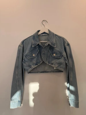 Cropped ljusblå jeansjacka från Zara - Trendig cropped jeansjacka från Zara i ljusblå denim. Klassisk krage, långa ärmar och två bröstfickor med knappar. Snygg oversized look och cool, kort passform som passar perfekt till high waist jeans eller kjol.