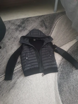 Moncler cardigan  - Snygg svart dunväst från Moncler med stickade ärmar och huva. Västen har dragkedja framtill, två fickor med tryckknappar och Moncler-logga på ena ärmen. Perfekt för lager-på-lager och streetstyle.
