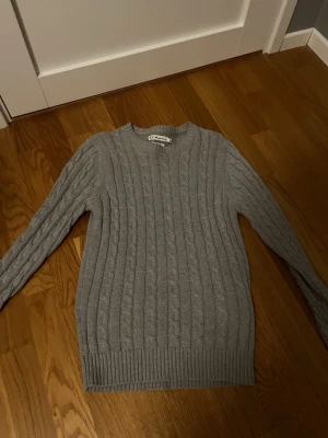 Grå kabelstickad tröja från S.C.W. - Snygg grå kabelstickad tröja från S.C.W. i återvunnet material. Tröjan har rund halsringning, ribbade muddar och klassiskt flätmönster. Perfekt för dig som gillar stilrena och bekväma plagg med en modern touch.