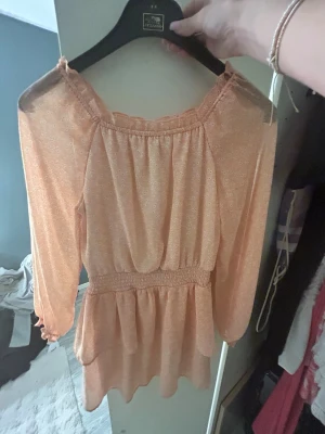 Persikofärgad blus från Kappahl - Supersöt persikofärgad blus från Kappahl med små vita blommor, smock i midjan och volangkant nertill. Blusen har långa ärmar och en avslappnad passform, perfekt för en trendig och bekväm look.