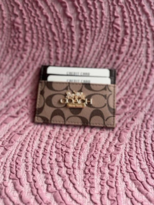 Coach brun kortfodral med logga - Snyggt kortfodral från Coach i brunt konstläder med det klassiska C-mönstret och guldfärgad logga framtill. Fodralet har flera fack för kort och är perfekt för dig som vill ha en stilren och kompakt plånbok.