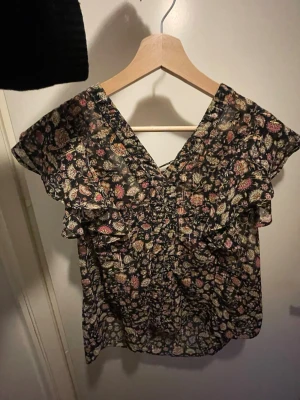 Blommig blus med volang och snörning - Säljer en blommig blus med volangdetaljer och snörning i ryggen. Blusen har v-ringad hals och korta ärmar. Färgerna går i svart, rosa, beige och grön. Perfekt för dig som gillar romantiska och boho-inspirerade plagg.