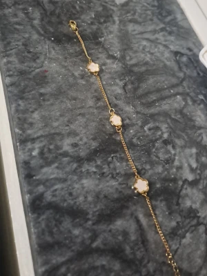 Guldfärgat armband med vita blommor - Snyggt armband i guldfärg med tre vita fyrklöverformade detaljer. Kedjan är tunn och elegant, perfekt för att addera lite stil till din outfit. Passar dig som gillar minimalistiska och trendiga accessoarer.