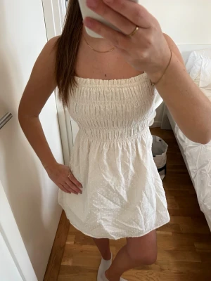 Vit ärmlös kortklänning med smock - Söt vit kortklänning med smock upptill och lätt utställd kjol. Klänningen är ärmlös och har en diskret prickig struktur i tyget. Perfekt för varma dagar och enkel att styla med accessoarer.