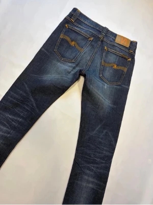 Mörkblå Nudie Jeans - Snygga mörkblå jeans från Nudie Jeans med klassisk femficksdesign och orangea sömmar. Jeansen har en slim fit-modell och är tillverkade i denim med lätt slitna detaljer och Nudie-logga på bakfickan. Hör av dig vid frågor elr likande!