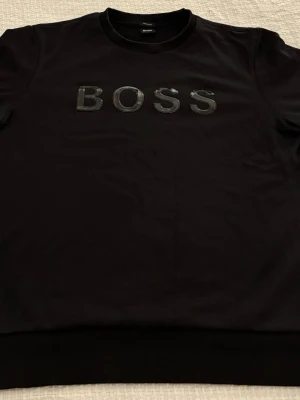 Svart Hugo Boss sweatshirt - Hugo Boss sweatshirt i svart.  Stilren crewneck-tröja från Hugo Boss med diskret gummerad BOSS-logga på bröstet. Minimalistisk och exklusiv design som passar både till vardags och mer uppklädda outfits. Loggan har en lätt glans och kan skina i ljus.  • Storlek: XL (se bild på etikett) • Passform: Regular fit • Färg: Svart • Material: Mjuk och följsam kvalitet • Detaljer: Rund hals, mudd vid ärmslut och nederkant  Skick: Mycket bra skick – endast använd några få gånger. Inga fläckar eller hål.
