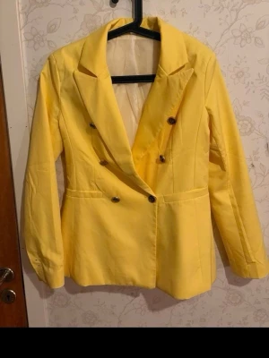 Gul dubbelknäppt kavaj - Snygg gul kavaj med dubbelknäppning och svarta knappar. Klassisk krage och två fickor framtill. Kavajen har en rak passform och är fodrad på insidan. Perfekt för dig som vill sticka ut med färg. Materialet känns som bomull eller polyester.