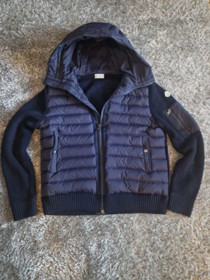 Mörkblå dunväst från Moncler - Snygg mörkblå dunväst från Moncler med stickade ärmar och huva. Västen har två fickor med knappstängning och dragkedja framtill. Klassisk Moncler-logga på ärmen och ribbade muddar för extra komfort. Perfekt för lager-på-lager och streetstyle.