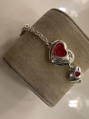 Guess armband med röda hjärtan - Snyggt armband från Guess i silverfärgad metall med två hjärtformade hängen dekorerade med röda detaljer och ett litet Guess-logohänge. Perfekt för alla hjärtans dag!😍