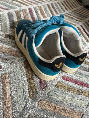 Blå Adidas Originals sneakers Campus - Snygga blå Adidas Originals sneakers med klassiska vita ränder på sidorna och guldfärgad logga på plösen. Skorna har en låg profil, vita sulor och svart detalj med gul Adidas-logga på hälen. Ovandelen är i mocka och textil för en retrovibe.