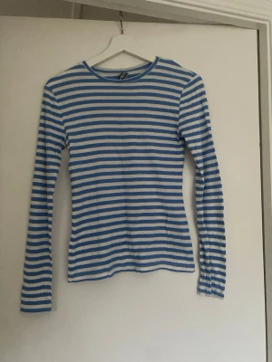 Blåvit randig långärmad topp - Säljer en blåvit långärmad topp med smala horisontella ränder. Toppen har rund halsringning och är ribbad för en skön passform. Perfekt att styla med jeans eller kjol för en enkel och fräsch look.