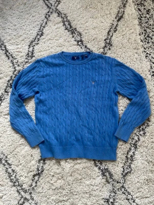 Blå kabelstickad tröja från Gant - Kabelstickad blå tröja från Gant med rund halsringning och diskret logga på bröstet. Tröjan har ribbade muddar vid ärmslut och nederkant, perfekt för en stilren och klassisk look.