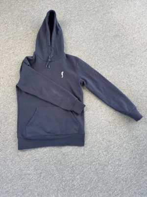 RS Sports Hoodie - Mörkblå hoodie med huva och dragsko, samt en liten vit logga på bröstet. Klassisk känguruficka framtill och ribbade muddar vid ärmslut och nederkant. Perfekt för en avslappnad och stilren look.