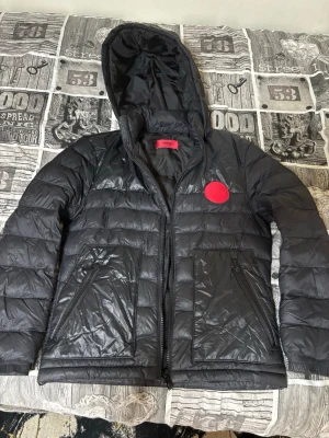 Hugo boss jacka - Snygg svart dunjacka från Hugo med huva och dragkedja framtill. Jackan har två stora fickor med dragkedja och en röd Hugo-logga på bröstet. Perfekt för kalla vinterdagar och har en modern, sportig look.