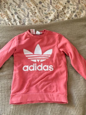 Rosa Adidas sweatshirt med logga - Cool rosa sweatshirt från Adidas med stor vit Trefoil-logga och text på bröstet. Tröjan har rund hals, långa ärmar och ribbade muddar vid ärmslut och nederkant. Perfekt för en avslappnad och sportig stil.