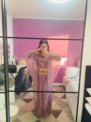 Kurdisk klänning  - Säljer en elegant långklänning i lila med glittrig spets och transparenta vida ärmar. Klänningen har ett dekorativt guldigt bälte i midjan och omlottkänsla framtill. Perfekt för dig som vill sticka ut med en unik look.