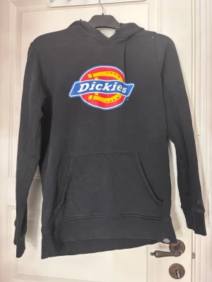 Svart Dickies hoodie med logga - Svart hoodie från Dickies med stor färgglad logga på bröstet. Klassisk känguruficka framtill. Perfekt för en avslappnad streetwear-look. Mjuk bomullskänsla och ribbade muddar vid ärmslut och nederkant. Finns någon fläck samt att kraken har gått sönder lite 