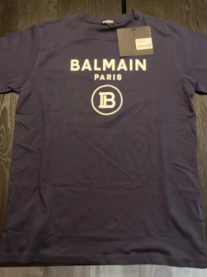 Mörkblå Balmain Paris t-shirt - Snygg mörkblå t-shirt från Balmain Paris med vit logotyptryck framtill. Klassisk rund halsringning och korta ärmar. Perfekt för dig som vill ha en stilren och trendig look med exklusiv känsla.