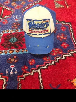 Blå och vit truckerkeps Western - Säljer en blå och vit truckerkeps med mesh på sidorna och böjd skärm. Framtill finns ett stort broderat märke med texten 'Western New York Vintage Adventures Begin'. Kepsen har slitna detaljer för en vintagekänsla.