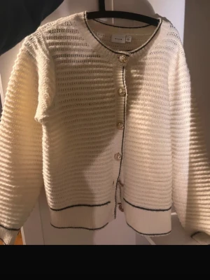 Vit kofta - Säljer en krämvit stickad kofta från Vila med dekorativa svarta kanter och guldfärgade knappar framtill. Koftan har rund halsringning, lång ärm och ribbade muddar. Perfekt för dig som gillar en clean och stilren look. jag vill påpeka att jag har sytt vissa knappar som har åkt av. 