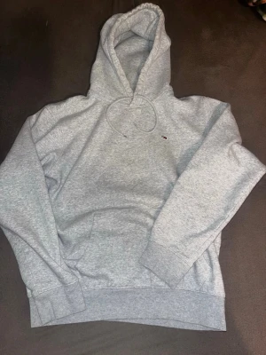 Grå hoodie från Tommy Hilfiger - Snygg ljusgrå hoodie från Tommy Hilfiger med liten broderad logga på bröstet. Klassisk passform med känguruficka, ribbade muddar och justerbar huva med dragsko. Perfekt för en chill och stilren look.