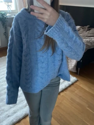Blå kabelstickad tröja - Mysig blå kabelstickad tröja med rund hals och långa ärmar. Tröjan har ett klassiskt flätmönster både fram och på ärmarna samt ribbade muddar vid hals, ärmslut och nederkant. Perfekt för kalla dagar och enkel att styla till olika outfits. ❤️