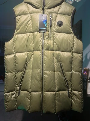 Philip plein väst  - Snygg olivgrön dunväst från Philipp Plein med huva och quiltad design. Västen har två snedställda dragkedjefickor fram, en bröstficka med dragkedja och ett svart logomärke på bröstet. Perfekt för lager-på-lager och streetwear. Ny pris 3900