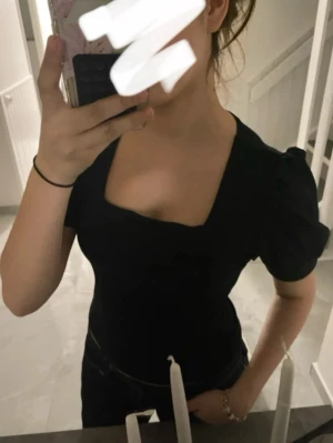 Svart topp med puffärm och fyrkantig ringning - Snygg svart topp med puffärmar och en trendig fyrkantig ringning. Modellen är figurnära och passar perfekt till jeans eller kjol. Toppens material känns mjukt och stretchigt, vilket gör den bekväm att bära hela dagen.