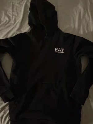 Svart hoodie från EA7 Emporio Armani - Säljer en svart hoodie från EA7 Emporio Armani med vit logga på bröstet. Klassisk modell med huva och magficka, tillverkad i mjukt material som känns skönt mot huden. Perfekt för dig som gillar stilrena och exklusiva plagg.