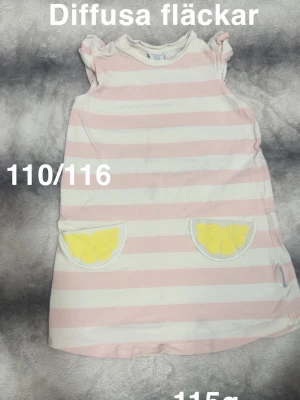 Randig rosa och vit klänning citron - Söt kortklänning med rosa och vita ränder samt två fickor med citronmotiv framtill. Klänningen har små volangärmarä, perfekt för varma dagar. Passar dig som gillar lekfulla detaljer och pastellfärger.🌸Diffusa fläckar. Storlek 110/116