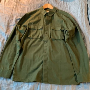 Grön overshirt från Pier One - Snygg militärgrön overshirt från Pier One med klassisk krage, två stora bröstfickor med lock och tryckknappar framtill. Skjortan är långärmad och tillverkad i bomull, perfekt för lager-på-lager-stil. Och helt ny har varit för stor på mig, så har inte kunnat haft på mig den.