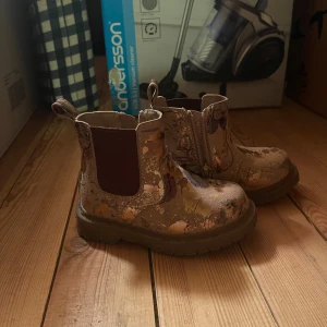 Blommiga chelsea boots med dragkedja - Säljer ett par snygga chelsea boots med blommigt mönster i beige och bruna toner. Skorna har elastiska paneler på sidorna, dragkedja på insidan och grov platt sula. Perfekta för dig som vill sticka ut med coola detaljer och bekväm passform.