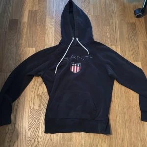 Svart hoodie från GANT med emblem - Svart hoodie från GANT med vit snörning i huvan och stort broderat GANT-tryck samt sköldemblem på bröstet. Klassisk känguruficka framtill och långa ärmar. Perfekt för en chill och sportig look.