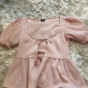 Ljusrosa blus med puffärm från Gina Tricot - Supersöt ljusrosa blus från Gina Tricot med puffiga korta ärmar och två knytningar framtill. Blusen har en lätt rynkad struktur och en luftig, flowy passform. Perfekt för dig som gillar romantiska detaljer och en feminin look.
