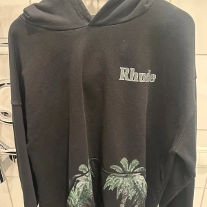 Svart hoodie Rhude med palmmotiv - Svart hoodie från Rhude med tryckt logga på bröstet och stort färgglatt motiv med palmer och figur på ryggen. Tröjan har huva och långa ärmar, perfekt för en avslappnad streetwear-look.