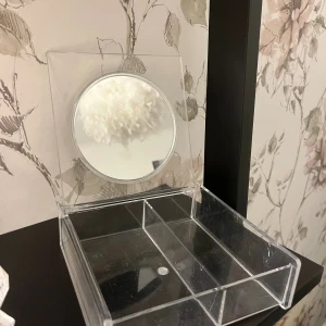 Transparent sminkspegel med förvaring - Säljer en stilren och transparent sminkspegel med inbyggd förvaringslåda i akryl. Spegeln är rund och sitter på ett lock som kan fällas upp. Perfekt för att organisera sminkborstar, läppstift eller andra små produkter.