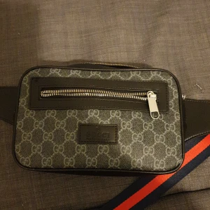 Gucci axelväska med GG-mönster - Snygg axelväska från Gucci med klassiskt GG-mönster i grått och svart. Använd 3 gånger så den är som Ny, Väskan har silverfärgade dragkedjor, svarta skinn-detaljer och en justerbar rem i marinblått och rött. Perfekt storlek för det viktigaste och riktigt stilren design.
