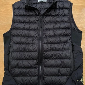 Svart dunväst från Stone Island (medium) - Säljer en svart dunväst från Stone Island med quiltad design och dragkedja framtill. Västen har två fickor med dragkedja och klassisk logotyp på sidan. Tillverkad i syntetmaterial och dun, perfekt för lager-på-lager-stil. Storleken är medium.