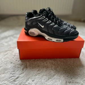 Nike Air Max Plus Tns Svarta Sneakers - Nike Air Max Plus Tn sneakers i svart med vita detaljer och ikoniska Tn Air-loggan på hälen. Ovandel i mesh med vågformade linjer och synlig Air Max-dämpning i sulan. Skorna har snörning och en sportig vibe som sticker ut. Perfekta för dig som gillar streetwear och snygg design. 