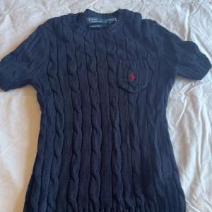 Mörkblå stickad tröja Polo Ralph Lauren - Säljer en mörkblå kabelstickad tröja från Polo Ralph Lauren i pima bomull. Tröjan har korta ärmar, rund halsringning och en liten bröstficka med röd broderad logga. Perfekt för dig som gillar klassisk och stilren look.