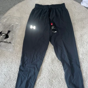 Svarta träningsbyxor från Under Armour - Svarta träningsbyxor från Under Armour med elastisk midja och snörning. Byxorna har en smal passform, reflekterande logga på benet och är tillverkade i lätt syntetmaterial som andas. Perfekta för träning och sportiga dagar. 