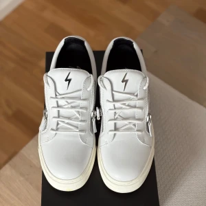 Vita sneakers från Giuseppe Zanotti - Snygga vita sneakers i skinn från Giuseppe Zanotti med svarta detaljer på hälen, silverfärgad blixt på plösen och dragkedjor på sidorna. Klassisk rund tå, vita snören och platt sula. Perfekta för dig som gillar stilrena men ändå unika skor.