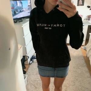 Svart hoodie från Tommy Hilfiger - Svart hoodie från Tommy Hilfiger med logga och text på bröstet. Klassisk huva med snörning och stor magficka framtill. Mjuk och skön passform, perfekt för chill dagar. Snyggt och stilrent plagg som funkar till jeans eller kjol.