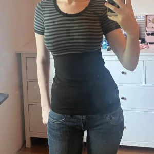 Randig svart och grå topp med spets - Säljer en slim, kortärmad topp med svart och gråa horisontella ränder upptill och enfärgat svart nedtill. Toppen har rund halsringning och små spetsdetaljer vid hals och ärmslut. Perfekt till jeans eller kjol för en chill look.