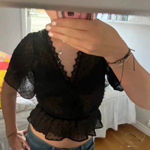 Svart blus med volang och spets - Säljer en svart transparent blus med prickigt mönster, v-ringning med spetskant och volang nertill. Blusen har korta, luftiga ärmar och markerad midja. Perfekt att styla med jeans eller kjol för en trendig look.