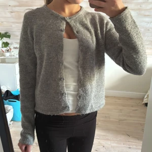 Grå stickad kofta med knappar - Mysig grå stickad kofta med rund hals och knäppning framtill.går att styla lite hur man vill och den har långa ärmar, perfekt att slänga över en topp. Enkel och stilren design som funkar till det mesta.