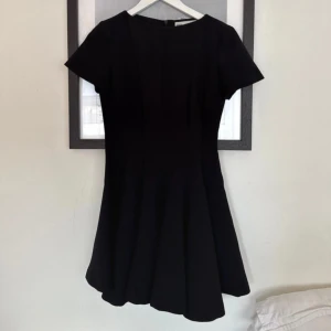 Svart kort klänning från Zara XS - En svart, kort klänning från Zara i storlek XS. Klänningen har rund hals, korta ärmar och en utsvängd kjol som ger en snygg siluett. Tillverkad i ett mjukt och följsamt material, perfekt för dig som gillar stilrena och enkla plagg.